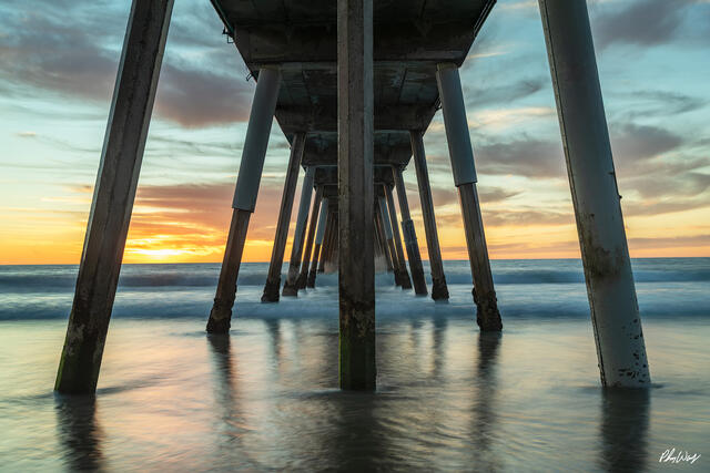 Sunset view beneath the Manhattan Beach pier with smooth ocean reflections and glowing horizon in California.