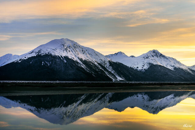 Snow covered mountains reflected in calm water beneath a soft golden sky in Kenai Alaska.