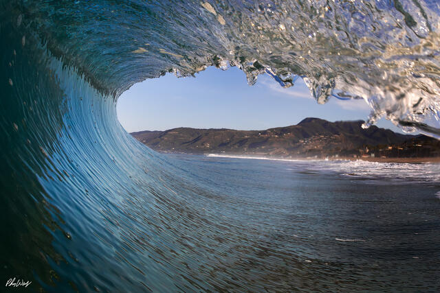 Ocean wave curling at Zuma Beach with Malibu coastline visible through the barrel in California.