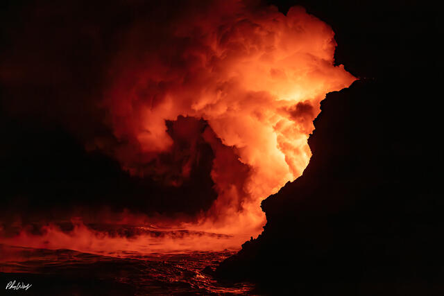 Tiki statue on rocky Hawaiian shoreline with palm trees under a glowing red volcanic sky at night.