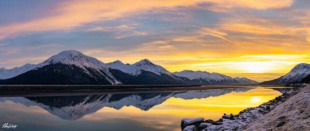 Snow covered mountains reflected in calm water at sunset with golden clouds in Kenai Alaska.
