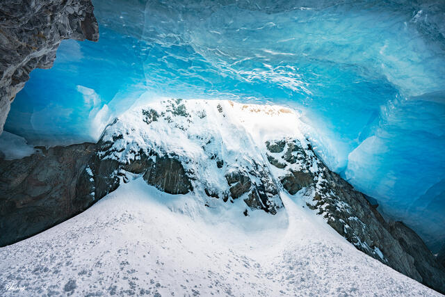 Blue ice cave interior with snow covered ground and exposed rock in a glacial landscape in Kenai Alaska.