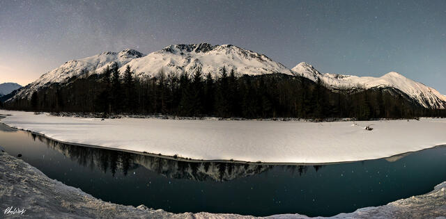 Snow covered mountains and a calm river beneath a star filled night sky in Kenai Alaska.
