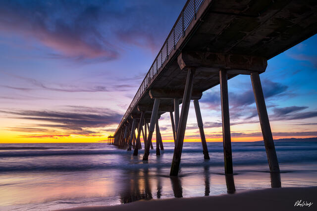 Manhattan Beach pier at sunset with purple and blue sky reflections over smooth ocean water in California.