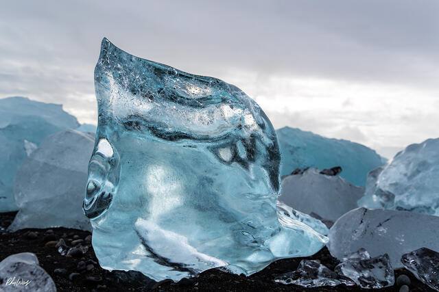 Blue glacier ice standing on black volcanic sand with scattered ice fragments and distant glaciers in Iceland.