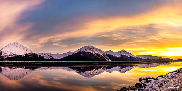 Snow covered mountain range reflected in calm water beneath a golden sunset sky in Kenai Alaska.