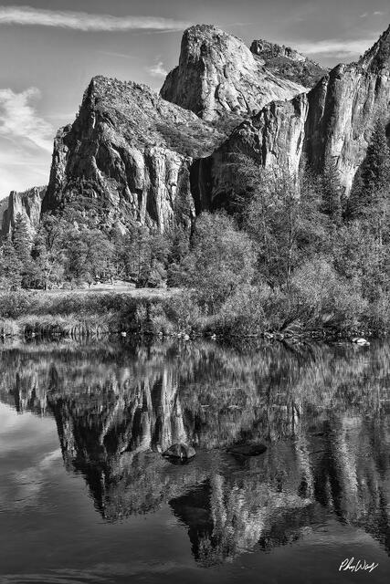 Granite cliffs and trees reflected in calm river water beneath rocky formations in black and white.
