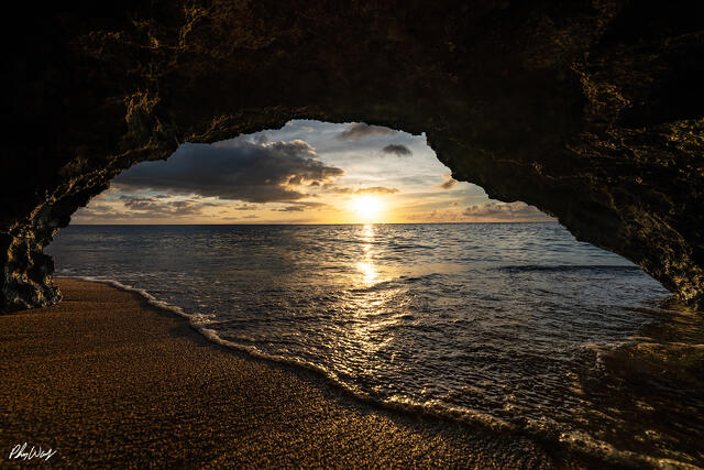 Sunset over ocean water viewed through a volcanic rock arch with waves reaching a sandy shoreline