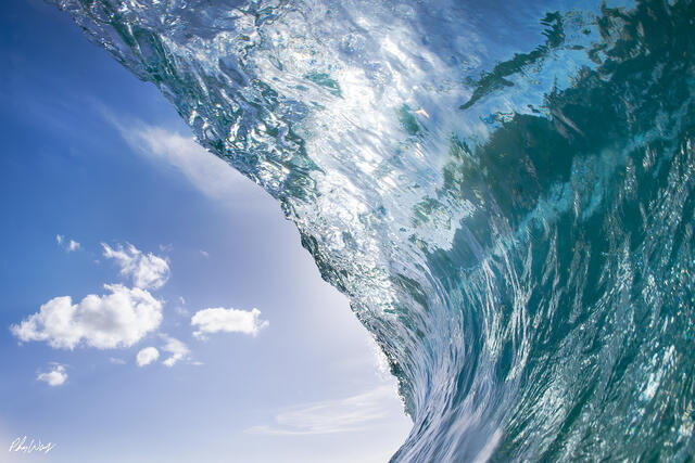 Close up view of a powerful ocean wave forming a translucent wall of water with blue sky and clouds above in Haleiwa, Oahu.