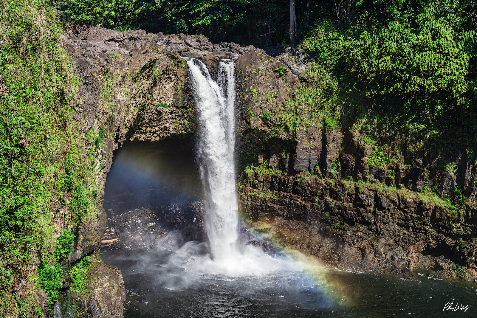 Rainbow Falls Rainbow Waterfall flowing into pool with rainbow in mist surrounded by tropical greenery in Hilo Hawaii.
