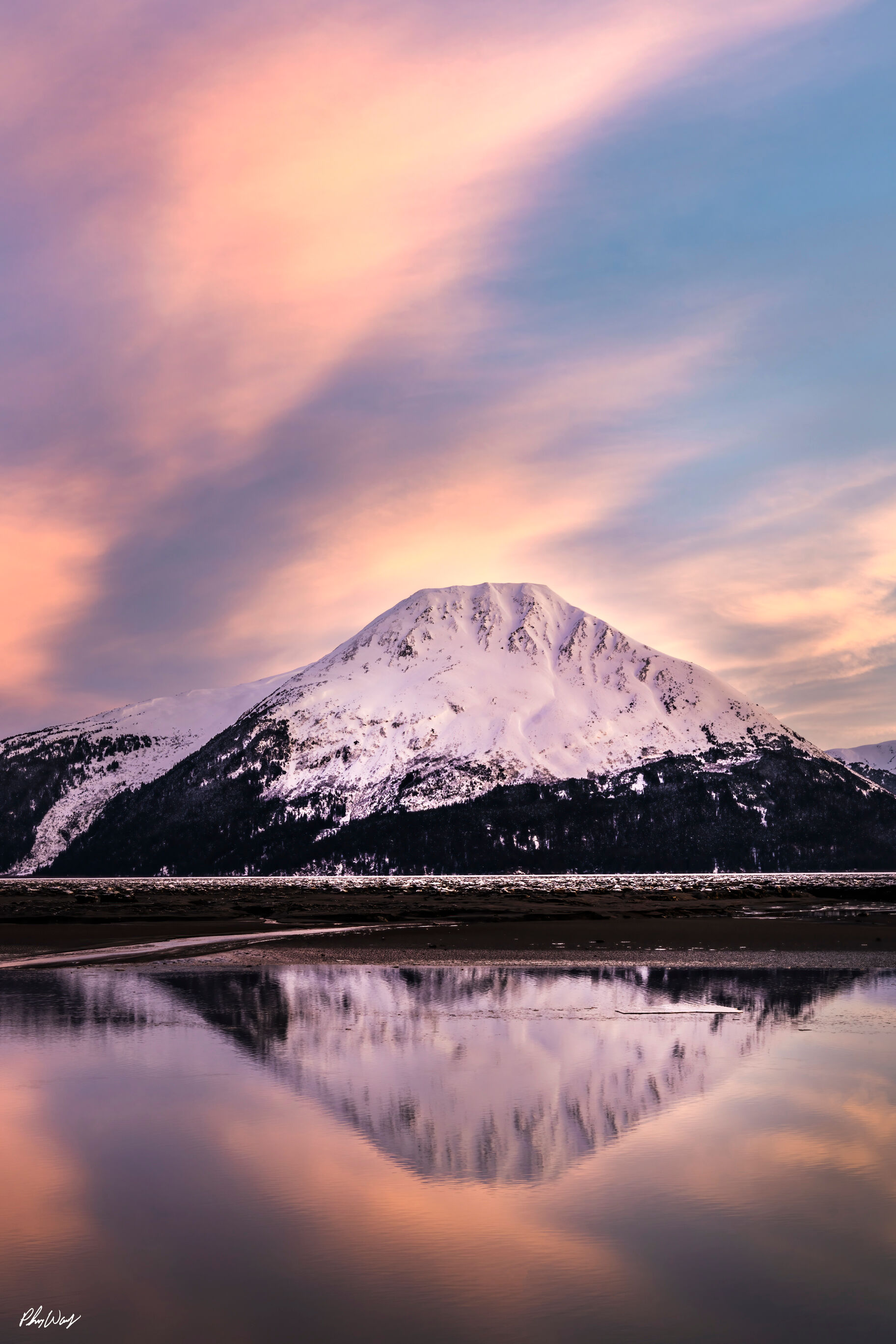 The Peak Snow covered mountain reflected in calm water beneath a pastel sunset sky in Kenai Alaska.